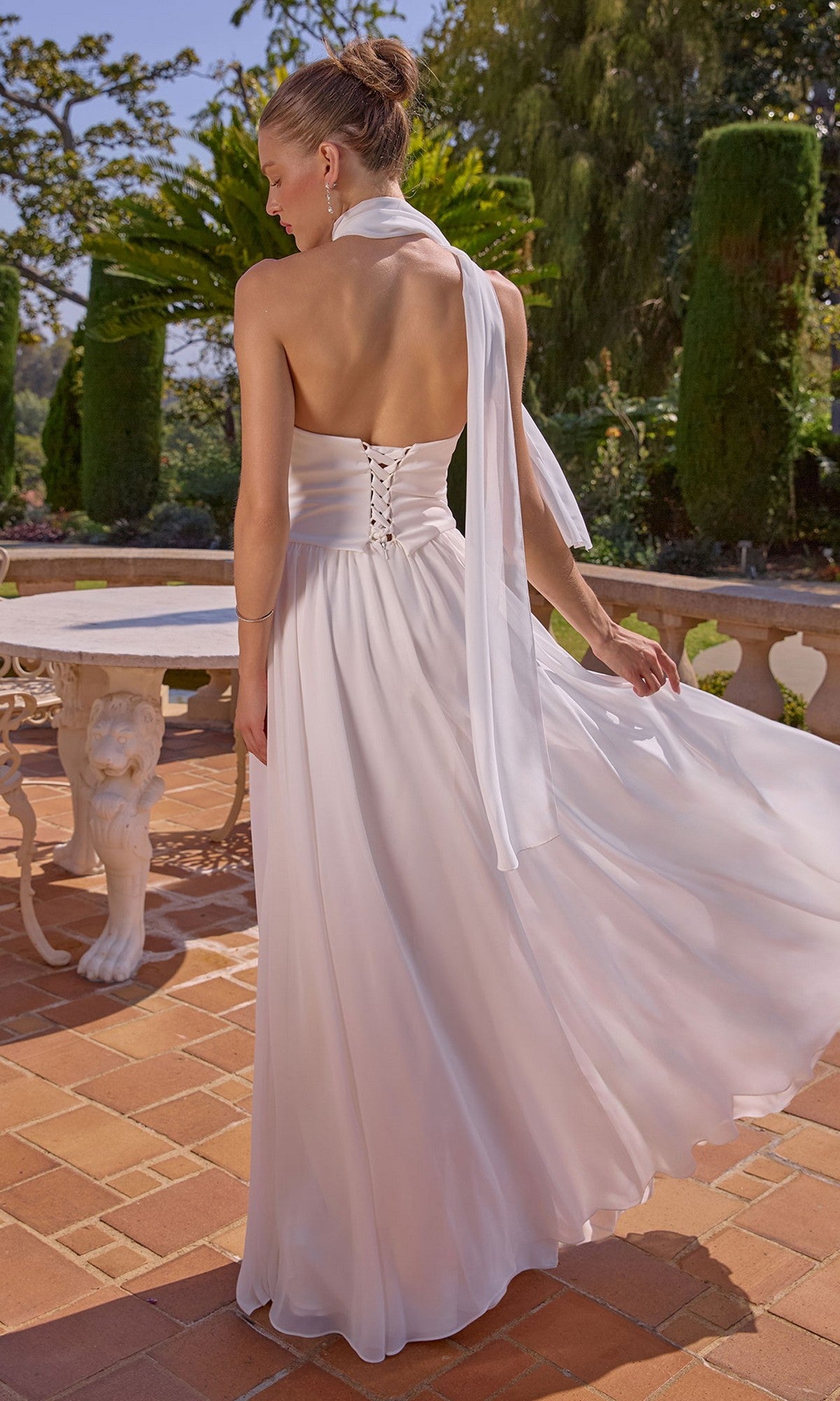 A woman wearing the Amoris T741W Long Prom Dress, with an off-white chiffon A-line skirt and lace-up back, stands on a tiled patio near a round table, surrounded by greenery and sunlight.